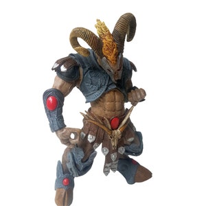 May include: A detailed action figure of a horned warrior with a red and blue armor, holding a weapon. The figure has a brown and gold horned helmet and a red gem on its chest.