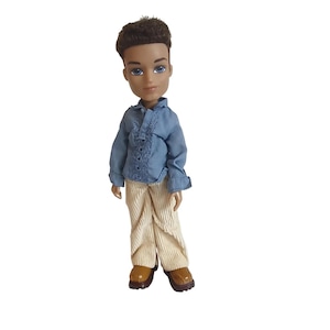 Könnte beinhalten: Eine Bratz-Puppe mit braunen Haaren und blauen Augen. Die Puppe trägt ein hellblaues Hemd mit Knöpfen, eine beigefarbene Cordhose und braune Schuhe. Die Puppe steht vor einem weißen Hintergrund.