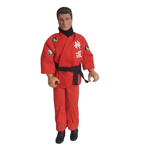 May include: A vintage action figure wearing a red karate uniform with a black belt. The uniform has white Japanese characters and patches on the sleeves. The figure has short brown hair and black shoes.