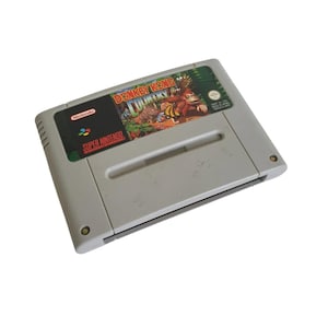 Op de afbeelding: Een grijze Super Nintendo videogame cartridge voor Donkey Kong Country. Het label bevat de speltitel in rode en gele tekst, een illustratie van Donkey Kong en Rambi de neushoorn in een jungleomgeving, en het Nintendo-logo.