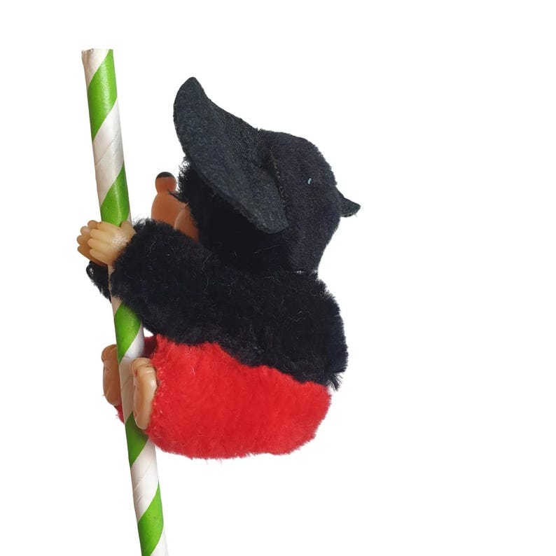 May include: A plush toy monkey with black fur, red pants, and a black hat climbs a green and white striped pole. The monkey's hands and feet are a light brown color.