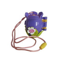 Puede incluir: Un juguete compacto Polly Pocket morado con forma de flor con un cordón rosa. El juguete tiene detalles en amarillo y verde, y una flor blanca con el logo de Polly Pocket. El cordón tiene una cuenta amarilla.
