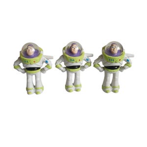 May include: Three Buzz Lightyear action figures. Each figure is white, green, and purple, with a clear helmet and wings. The figures are standing upright, with their arms at their sides. The toys are from the Toy Story franchise.