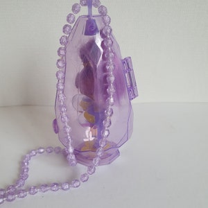 May include: A purple plastic doll charm with a beaded necklace. The doll is inside a clear plastic container shaped like a crystal.