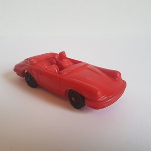 May include: A red toy car with a convertible top and two figures inside. The car has black wheels and a detailed front end.