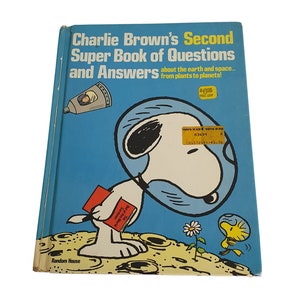 Puede incluir: Un ejemplar vintage de "Charlie Brown's Second Super Book of Questions and Answers" con Snoopy con un casco de astronauta en la portada. El libro tiene una cubierta azul con texto e ilustraciones.