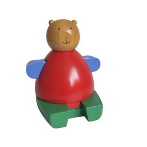 May include: A wooden bear toy with a light brown head, red body, blue arms, and green base. The bear has a simple face with a black smile and two small dots for eyes. The toy is designed for children.