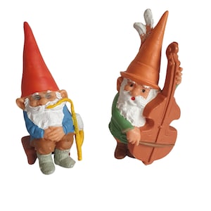 May include: Two small gnome figurines, one playing a cello and the other holding a pipe. The gnome playing the cello is wearing a green shirt and an orange hat. The gnome holding the pipe is wearing a blue shirt and a red hat.