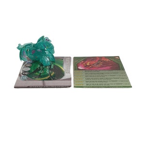 Puede incluir: Un juguete Bakugan verde y morado con una carta que dice "SUMMERGATE". La carta tiene un Bakugan rojo y negro y enumera las estadísticas del Bakugan: "POWER: 140", "G-POWER: 110", "ABILITY: 10", "DEFENSE: 180", "SPEED: 210", "ABILITY: 10".