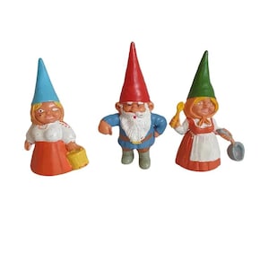 Op de afbeelding: Drie kleine gnoomfiguren. Eén gnoom draagt een rode hoed en blauwe kleding, een ander draagt een blauwe hoed en een rode jurk, en de derde draagt een groene hoed en een rode jurk.