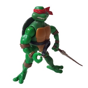 Könnte beinhalten: Grüne Teenage Mutant Ninja Turtles Actionfigur mit rotem Bandana, braunem Gürtel und einer braunen und silbernen Waffe. Die Figur ist in Kampfstellung.