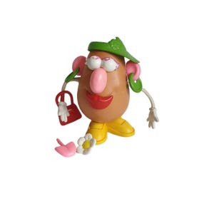 Puede incluir: Un juguete caprichoso de cabeza de patata con cuerpo marrón, nariz rosa y labios rojos. Lleva una visera verde, pendientes rosas y zapatos amarillos. Los accesorios incluyen un bolso rojo, una pala rosa y una flor blanca.