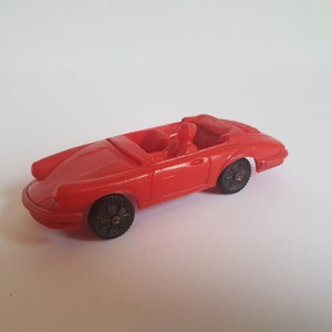 May include: A red toy car with a black interior and black wheels. The car is a convertible and has a driver figure inside.