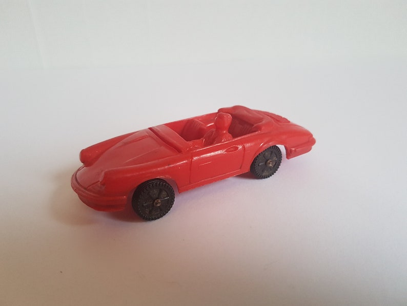 May include: A red toy car with a black interior and black wheels. The car is a convertible and has a driver figure inside.