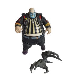Peut inclure: Figurine d'action détaillée d'un grand personnage chauve à la peau bleue et au sourire sinistre, portant une chemise rayée noire et jaune, un pantalon noir et une veste noire. Une figurine séparée, griffue, est également présente.
