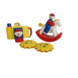 Op de afbeelding: Een verzameling vintage kinderspeelgoed. Bevat een gele speelgoedcamera met een rood-wit gezicht, een rood-wit schommelpaard met een blauwe ruiter en twee gele zonvormige speeltjes met lachende gezichten. Het speelgoed is gemaakt van plastic.
