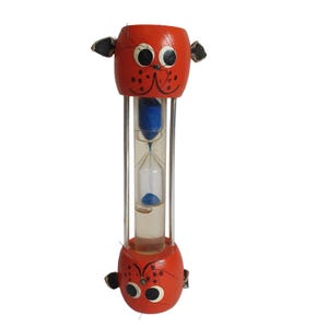 Peut inclure: Un sablier en bois orange fantaisiste avec un design de visage de dessin animé. Le sablier comporte un tube en verre transparent rempli de sable bleu, maintenu entre deux visages en bois orange avec des détails noirs et blancs. Le design comprend de petites oreilles noires.