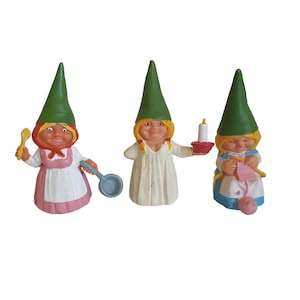 Puede incluir: Tres pequeñas figuras de gnomos con sombreros puntiagudos verdes. El gnomo de la izquierda sostiene una cuchara de madera y una sartén azul. El gnomo del medio sostiene un candelero con una vela blanca. El gnomo de la derecha sostiene una bola de lana rosa y agujas de tejer.