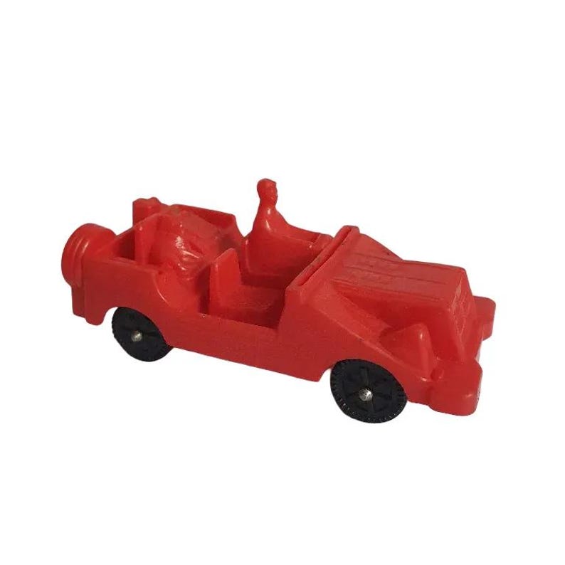 May include: A small, red toy car with black wheels. The toy car has a spare tire on the back and a driver figure. The car is a vintage style toy.
