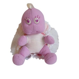 Peut inclure: Une tortue en peluche violette avec un tissu rayé rose et blanc sur son corps. La tortue a une bordure en dentelle blanche autour de sa carapace et de grands yeux roses.