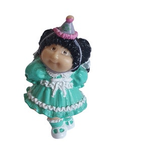 以下が含まれることがあります： 緑のレースの縁取りのあるドレスとピンクと紫のパーティーハットを着たキャベツパッチキッズのドールフィギュア。人形は黒髪で、顔はペイントされています。