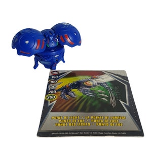 May include: Blue and red Bakugan toy with a card showing its stats and abilities. The card has the text "Point of Light - La Pointe de Lumiere - Punto de Luz - Punta di Luce - Punkt des Lichts - Ponto de Luz".