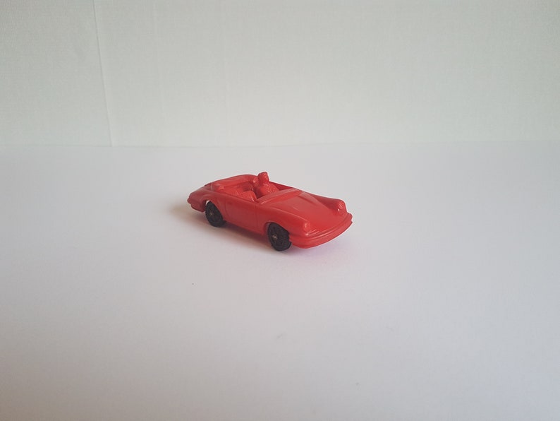 May include: A red toy car with a convertible top and two figures inside. The car has black wheels and is made of plastic.