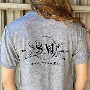 Puede incluir: Una camiseta gris jaspeada con un logotipo circular negro en la espalda. El logotipo presenta las letras "SM" dentro de un círculo, con diseños en forma de hojas y el texto "SAVVYMOCKS" debajo. La persona lleva vaqueros azules.