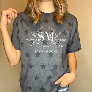 May include: A dark gray t-shirt with a white star pattern. The shirt has a white circle with the letters "SM" inside and the text "SAVVYMOCKS" below the circle.