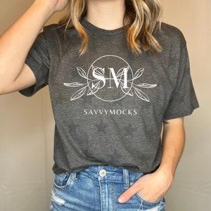 May include: A dark gray t-shirt with a white circle design featuring the letters "SM" inside. The circle is surrounded by white leaves and the text "SAVVYMOCKS" is printed below the circle.