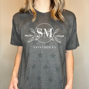 May include: A dark gray t-shirt with a white graphic design featuring the initials "SM" in a circle with leaves and the text "SAVVYMOCKS". The shirt has a subtle star pattern.