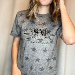 May include: A gray t-shirt with a star pattern and the text "SM SAVVYMOCKS" in a circular design. The shirt is short-sleeved and made of a soft material. The stars are a darker shade of gray.
