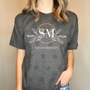 May include: A dark gray t-shirt with a white graphic design featuring the letters "SM" in a circle with leaves and the text "SAVVYMOCKS" below. The shirt has a relaxed fit and is made of a soft, comfortable fabric.