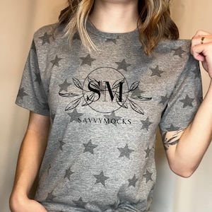 May include: A gray t-shirt with a star pattern and a black graphic design featuring the initials "SM" in a circle with leaves and the text "SAVVYMOCKS" underneath.