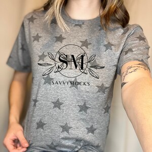 May include: A gray t-shirt with a white star pattern. The shirt has a black graphic with the letters "SM" inside a circle and the text "SAVVYMOCKS" below the circle.