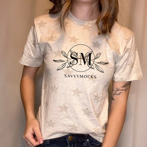 May include: Beige t-shirt with a star pattern and the text "SM SAVVYMOCKS" in a black circular design. The shirt is paired with dark blue denim jeans.