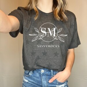 May include: Dark gray t-shirt with a white circular logo featuring the letters "SM" and the text "SAVVYMOCKS". The shirt is paired with blue denim shorts with distressed details.