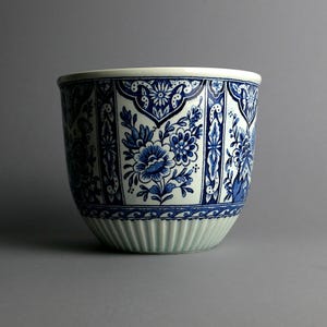 Op de afbeelding: Een keramische bloempot met een blauw en wit bloemenpatroon. De bloempot heeft een brede, afgeronde vorm met een witte rand en een geribbelde, lichtgroene basis. Het blauwe bloemendesign bevat bloemen en bladeren.