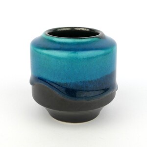 Peut inclure: Un petit récipient en céramique présentant une glaçure turquoise brillante et craquelée sur la partie supérieure, qui se transforme en une finition noire mate unie sur la section inférieure. Le bord est sombre et le corps est arrondi.