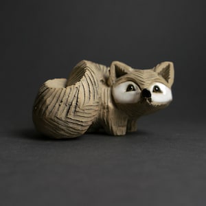 May include: A handcrafted ceramic raccoon figurine with a textured, carved design. The raccoon has white eyes, a black nose, and a curled tail that forms a small planter. The figurine is a neutral beige color, perfect for home decor.