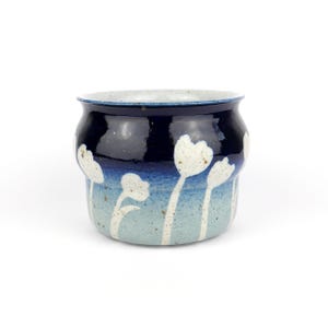 Pode incluir: Um vaso de cerâmica com uma borda azul escura e um gradiente de tons de azul. Flores brancas em forma de tulipa são pintadas na base azul clara. O vaso tem uma textura salpicada e uma forma arredondada.