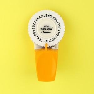 Könnte beinhalten: Ein weißes und orangefarbenes Mini Label-Mate-Etikettiergerät auf gelbem Hintergrund. Das Etikettiergerät hat ein kreisförmiges Zifferblatt mit Buchstaben und Zahlen sowie den Text "MINI LABEL-MATE by Dennison". Das Etikettiergerät ist orange und weiß.