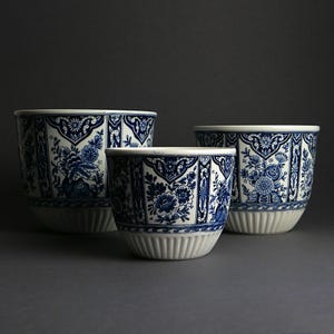 Könnte beinhalten: Drei blau-weiße Keramik-Blumentöpfe mit floralen Mustern. Die Töpfe haben einen weißen Rand und Boden mit vertikalen Rippendetails. Die blauen Designs umfassen Blumen und geometrische Formen. Die Töpfe sind in verschiedenen Größen erhältlich.