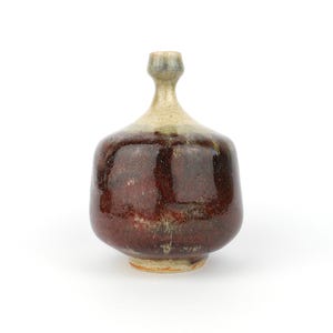 Può includere: Una bottiglia in ceramica fatta a mano con un corpo arrotondato e un collo sottile. La bottiglia presenta una ricca glassa lucida nei toni del rosso intenso e del marrone, con accenti beige chiaro e verdi vicino al collo. L'altezza è di circa 15 cm.