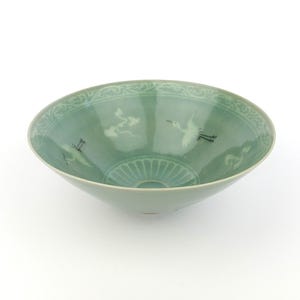 Peut inclure: Un bol en céramique vert céladon doté d'un bord large et évasé avec un motif de volutes en relief. La surface intérieure est décorée de deux illustrations de grues en vol et de motifs de nuages stylisés, avec un design cannelé à la base.