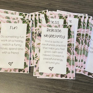 May include: A stack of pink and green cards with inspirational messages. The top card says "Release Negativity" and has a quote about mindfulness. The card below says "Fun" and has a list of activities to do for fun.