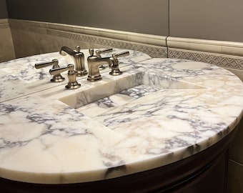Op de afbeelding: Een witte marmeren badkamer wastafel met twee wasbakken en een set van vier chromen kranen.