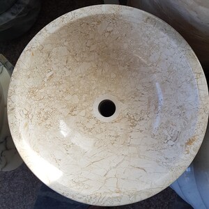 Pu&ograve; includere: Un lavandino in marmo bianco di forma circolare con un foro di scarico al centro. Il lavandino ha una superficie liscia e lucida con un sottile motivo a venature.