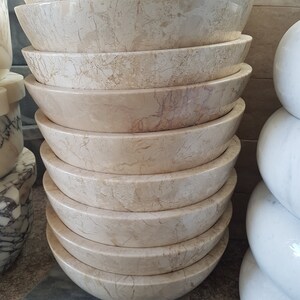 Pu&ograve; includere: Una pila di dieci ciotole rotonde in marmo beige con un motivo di venature marroni chiare. Le ciotole sono impilate a forma di piramide.