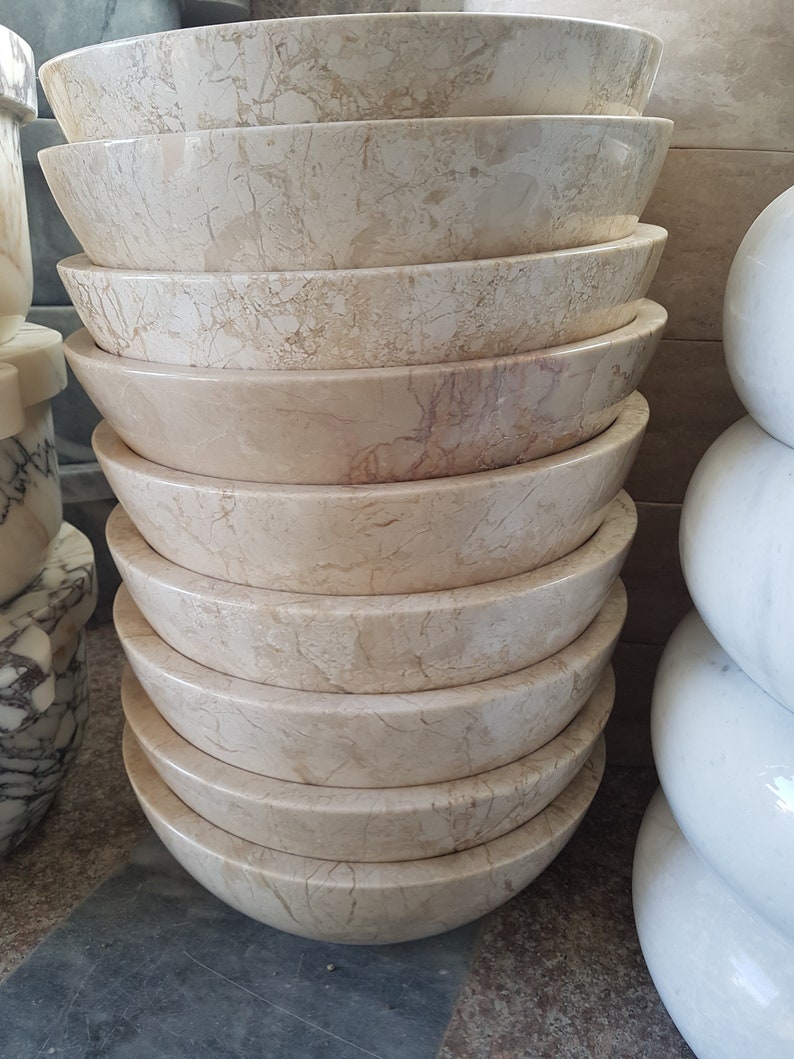 Pu&ograve; includere: Una pila di dieci ciotole rotonde in marmo beige con un motivo di venature marroni chiare. Le ciotole sono impilate a forma di piramide.